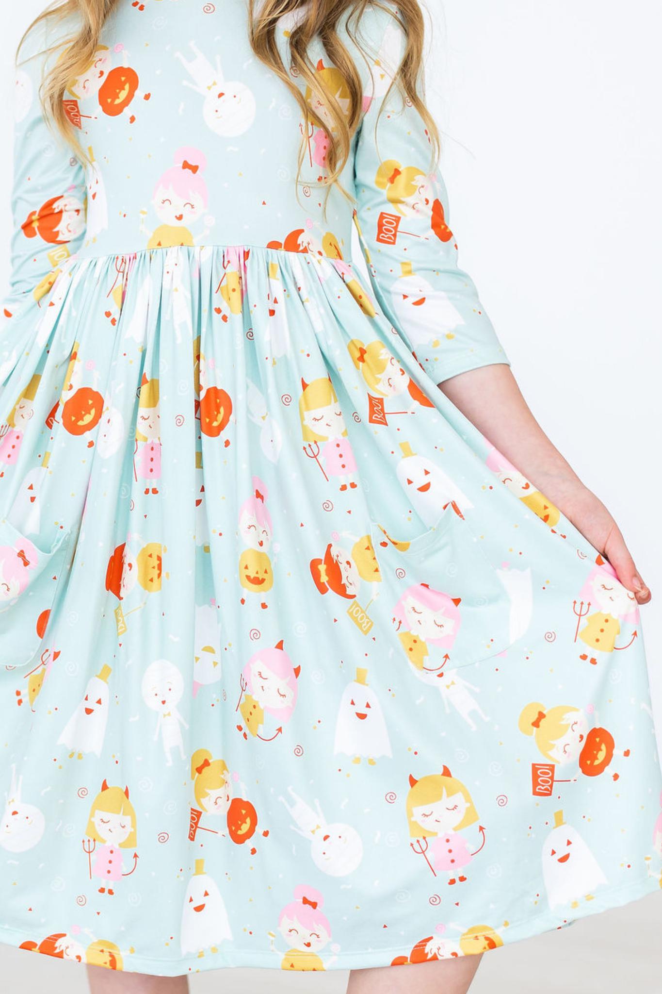 A child wearing a knee-length sleeve dress with a playful pattern featuring cartoon-like characters and Halloween-themed elements on a light blue background.