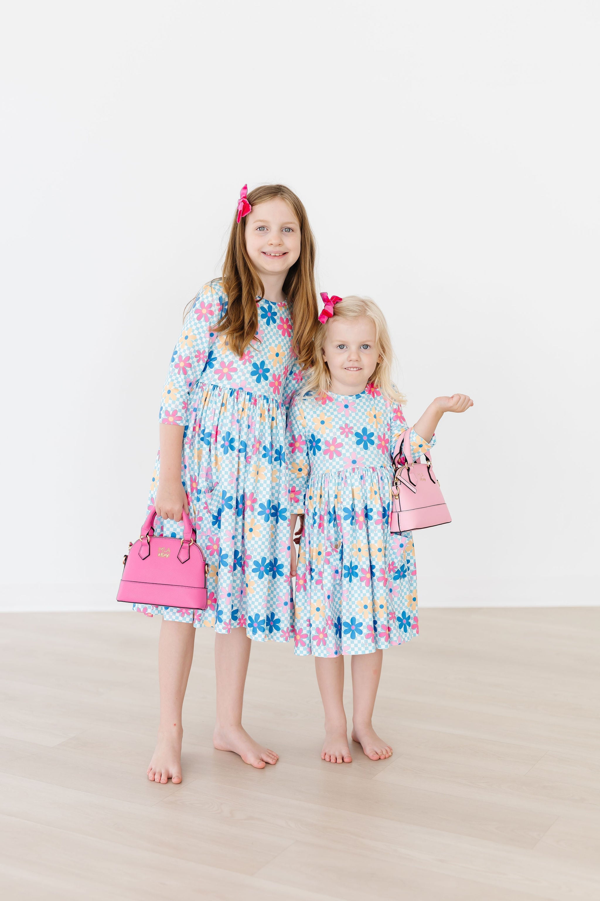 Two young girls wearing floral dresses with matching handbags on a plain background