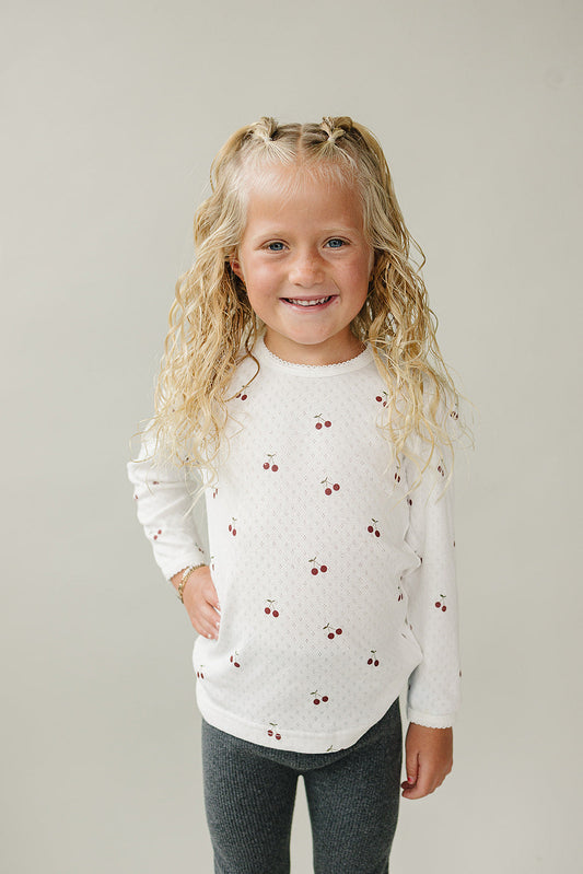 Young girl wearing a white long-sleeve shirt with red patterns against a plain background