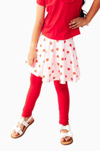 Child wearing a red top, white skirt with orange polka dots, and red leggings on a wooden floor.
